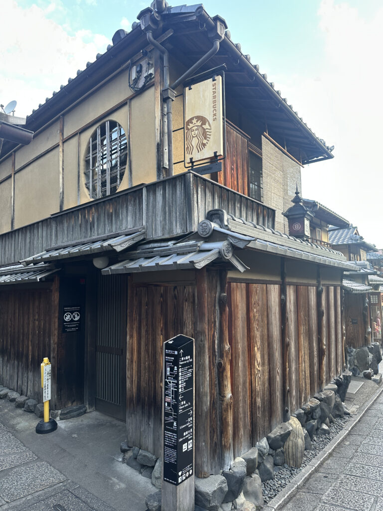 The image shows a traditional Japanese-style building with wooden walls and a tiled roof. A sign with the Starbucks logo is prominently displayed, indicating that the building houses a Starbucks store. The architecture features a circular window and wooden latticework, blending traditional Japanese design with modern branding. There are informational signs and a small barrier on the street in front of the building.