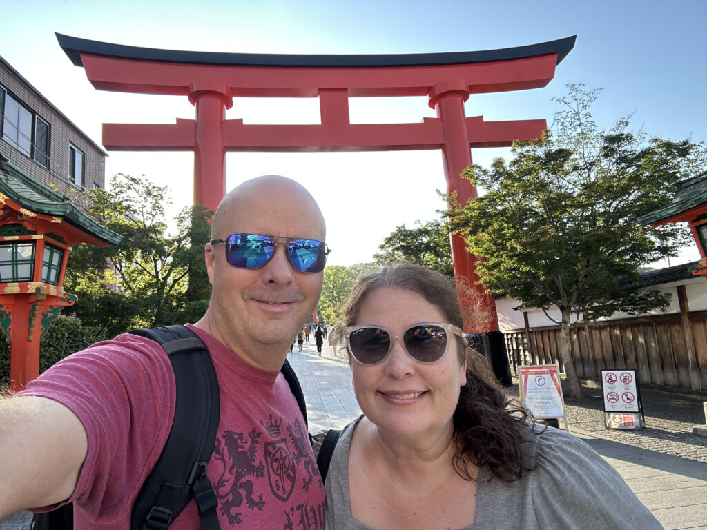 A man and a woman are taking a selfie in front of a large red torii gate, a traditional Japanese gate often found at the entrance of Shinto shrines. The man is wearing sunglasses and a red shirt, while the woman is also wearing sunglasses and a gray top. There are trees and a building in the background, and the sky is clear.