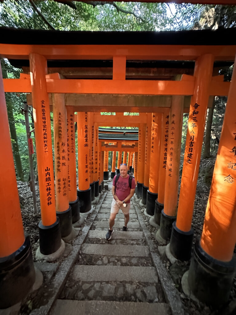 A person is walking up a stone staircase surrounded by a series of vibrant orange torii gates, which are inscribed with Japanese characters. The gates form a tunnel-like path, and the area is shaded by trees, creating a serene and picturesque setting.