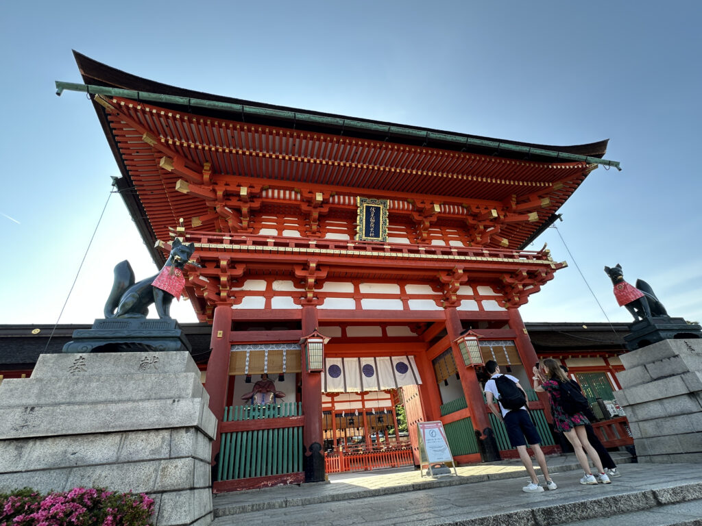 The image shows the entrance gate of a traditional Japanese shrine, characterized by its vibrant red and white colors. The gate is flanked by two fox statues, each wearing a red bib. Three people are standing in front of the gate, looking at the structure. The sky is clear and blue, providing a bright backdrop to the scene.