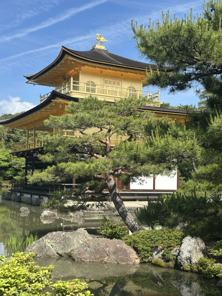 The image shows a traditional Japanese building with a golden exterior, known as Kinkaku-ji or the Golden Pavilion, surrounded by lush greenery and a pond. The structure has a distinctive roof with a golden ornament on top. The scene is serene, with clear blue skies and reflections in the water.