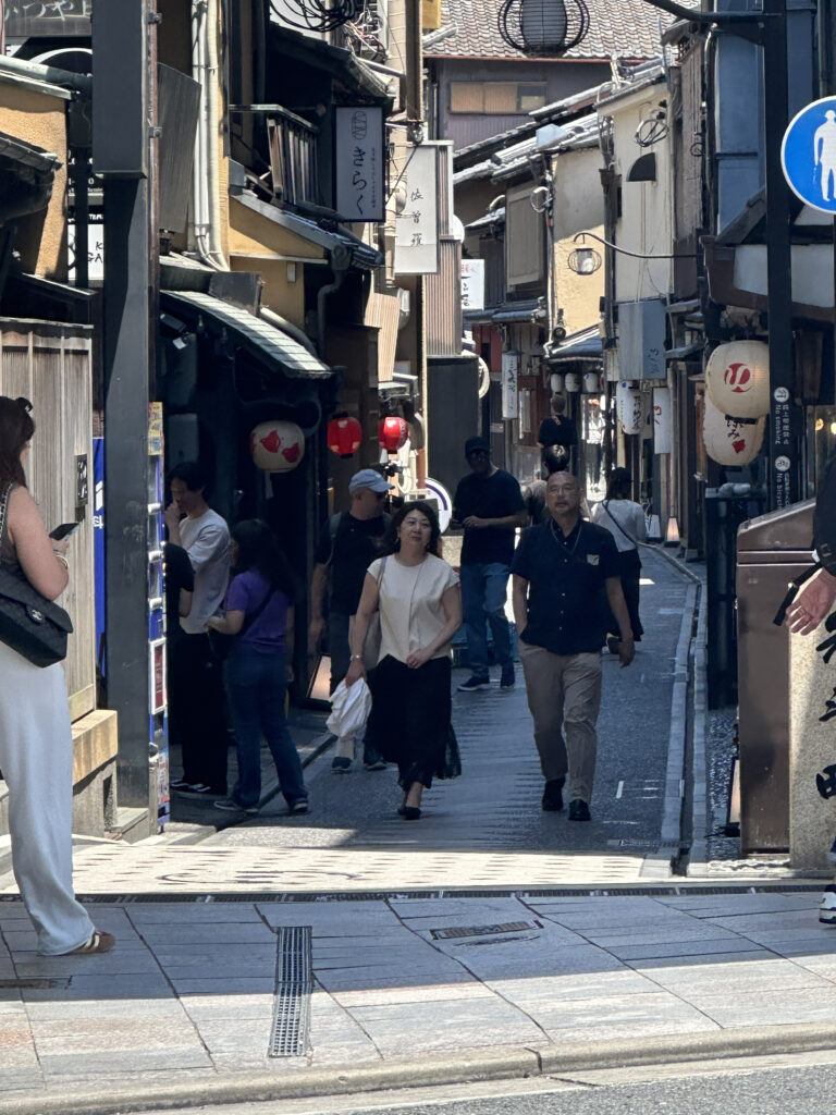 A narrow street in a traditional Japanese area, with several people walking. The street is lined with old-style buildings featuring wooden facades and hanging lanterns. Signs with Japanese characters are visible, and the atmosphere is bustling with pedestrians. The lighting suggests it is daytime.