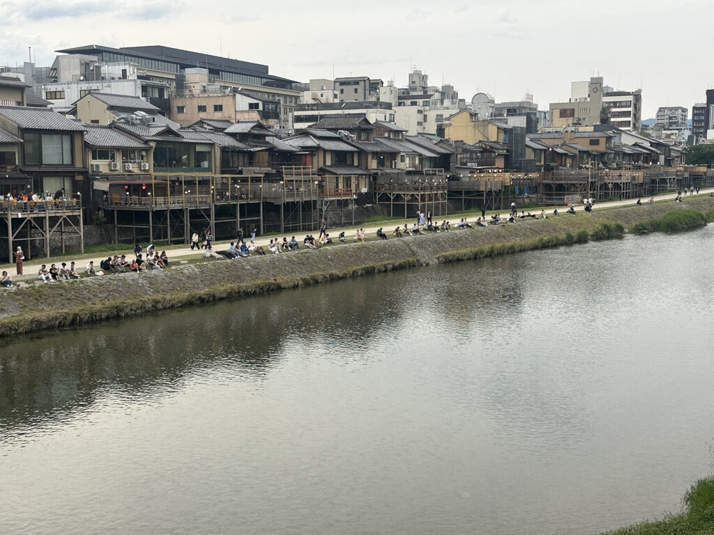 The image shows a riverside scene with people sitting along the grassy banks of a river. Across the river, there are traditional-style buildings with wooden balconies. The background features a mix of modern and older architecture, creating a contrast between the riverbank and the urban setting. The sky is overcast, adding a calm atmosphere to the scene.