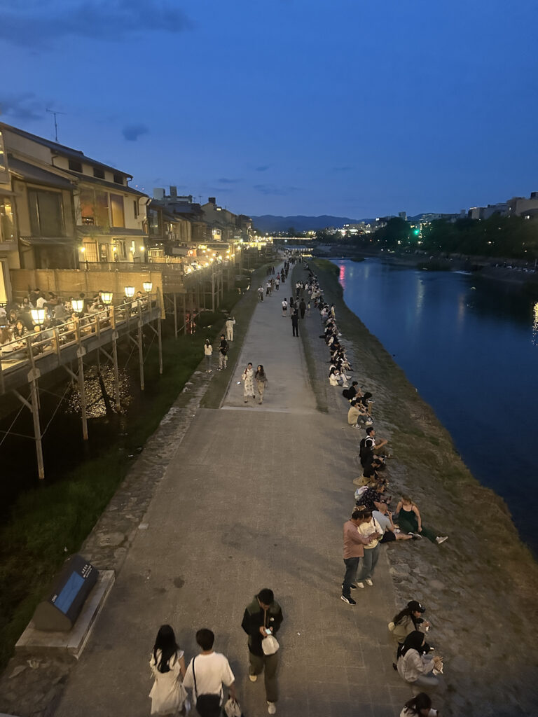 A scenic evening view of a riverside walkway with people sitting and strolling along the path. On the left, there are buildings with lit balconies, and on the right, a calm river reflects the evening sky. The scene is lively with groups of people enjoying the ambiance.