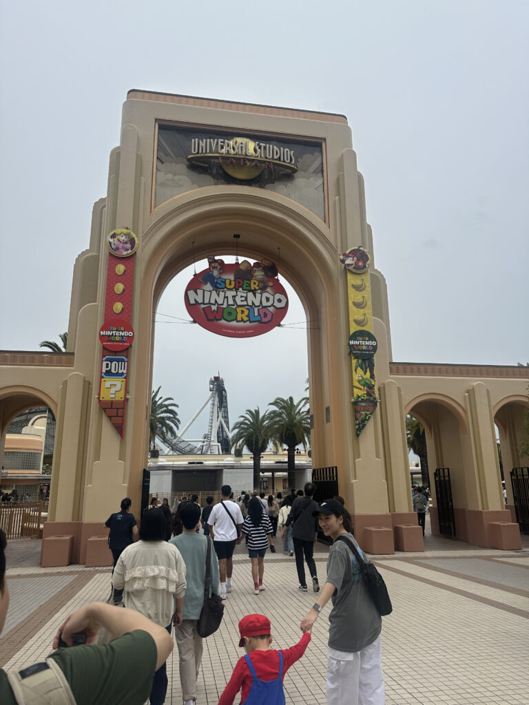 The image shows the entrance to Universal Studios Japan, featuring a large archway with a sign for "Super Nintendo World." The arch is decorated with various Nintendo-themed elements, including characters and symbols. People are walking towards the entrance, including a child dressed in a red and blue outfit resembling a popular video game character. Palm trees and a roller coaster are visible in the background.