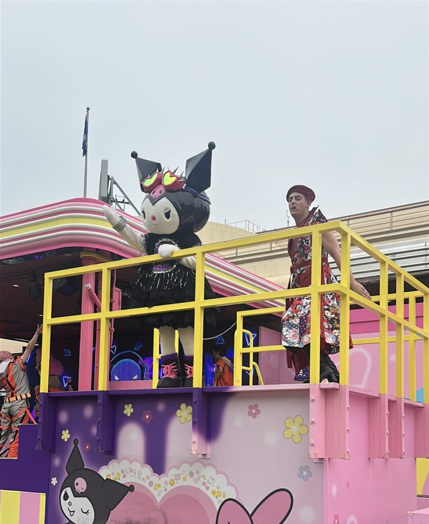 The image shows a colorful parade float decorated with Sanrio characters, including Kuromi and My Melody. A person in a vibrant outfit stands on the float next to a large Kuromi figure. The float is adorned with pink and yellow colors, flowers, and cartoon images. A child with a floral scrunchie in their hair is watching the parade.
