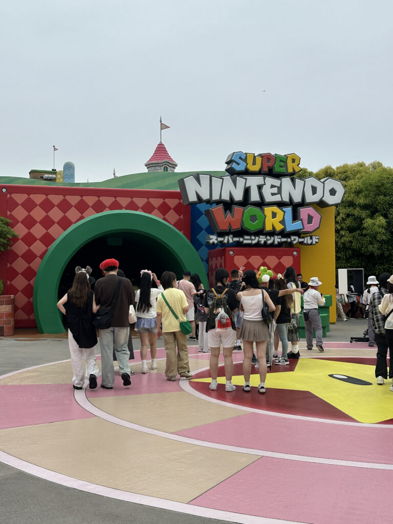 A group of people is gathered outside the entrance to "Super Nintendo World." The entrance features a colorful sign with the words "Super Nintendo World" in bold, playful letters. The area has a vibrant design with a large star pattern on the ground. The background includes a green hill with a castle-like structure and flags.