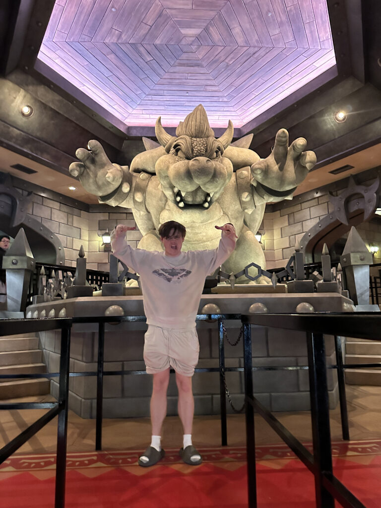 A person is standing in front of a large statue of a fictional creature with horns and a spiked shell, resembling a character from a video game. The setting appears to be indoors, with a decorative ceiling and stone-like walls. The person is wearing a light-colored outfit and is posing with their arms raised.