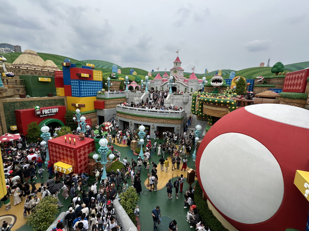 The image shows a vibrant theme park area inspired by a video game, featuring colorful structures resembling blocks and characters from the game. There are large, whimsical decorations like a giant red and white mushroom and a castle-like building. The area is bustling with many visitors walking around and exploring the attractions. The background includes green hills and various themed elements, creating an immersive environment.
