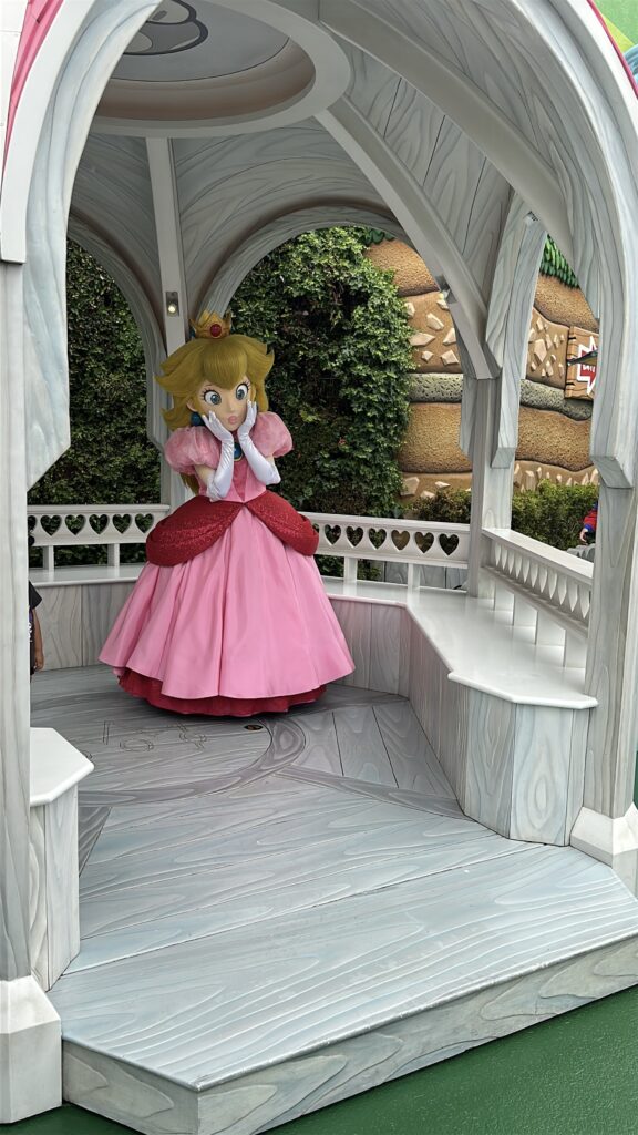 A person in a Princess Peach costume is standing inside a small, white gazebo. The costume features a pink dress with puffed sleeves, a red overlay, white gloves, and a crown. The gazebo has heart-shaped cutouts on the railing, and there is greenery and a stone wall in the background.