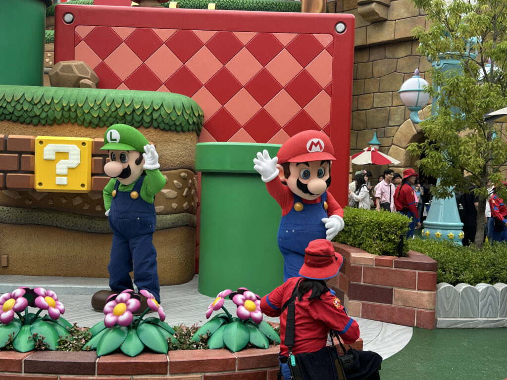 The image shows two people dressed in costumes of popular video game characters, one in a red outfit with a red hat and the other in a green outfit with a green hat. They are standing in a themed area resembling a video game environment, with elements like a large green pipe, a question mark block, and colorful flowers. A person in a red hat and jacket is taking a photo of them. There are other people and themed decorations in the background.