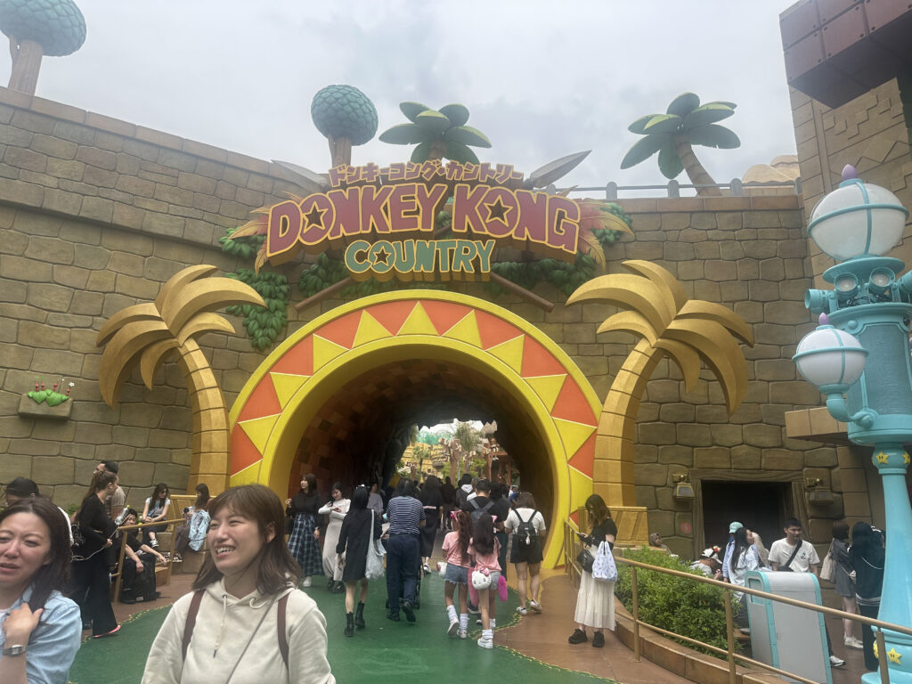 The image shows the entrance to "Donkey Kong Country" at a theme park. The entrance is designed with a large, colorful archway featuring palm tree decorations and a sign with the name "Donkey Kong Country" in bold letters. People are walking towards and away from the entrance, and the atmosphere appears lively and busy. The surrounding area includes themed decorations and structures.