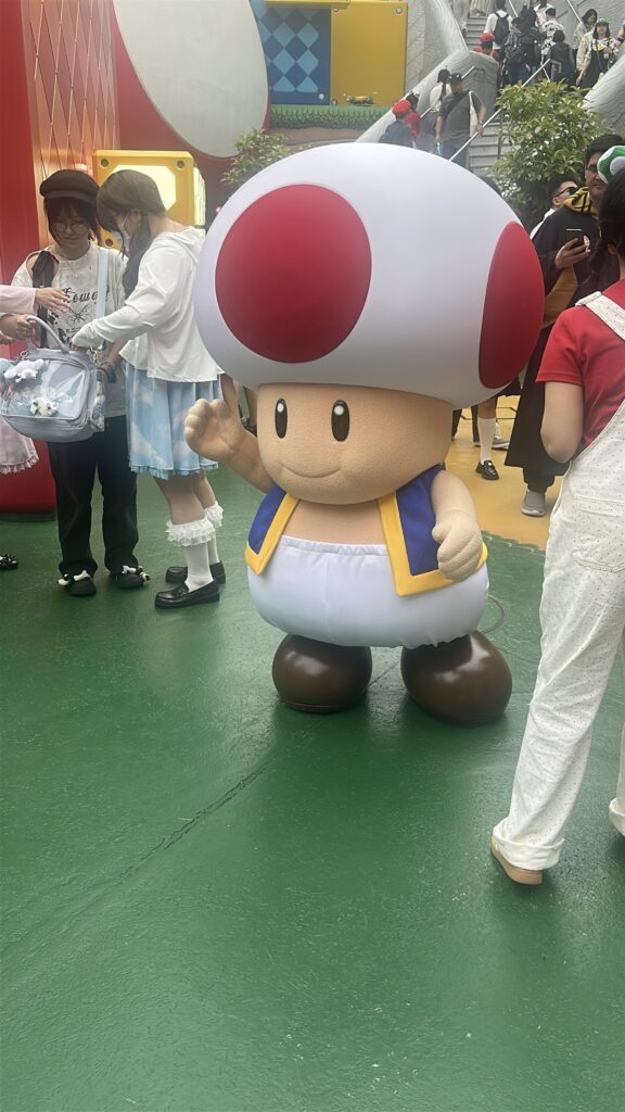 A person in a Toad costume from the Mario franchise is standing on a green floor. The costume features a large white mushroom cap with red spots, a beige face, and a blue and yellow vest. Several people are around, some interacting with the character. The background includes colorful, themed decor and a staircase with more people.