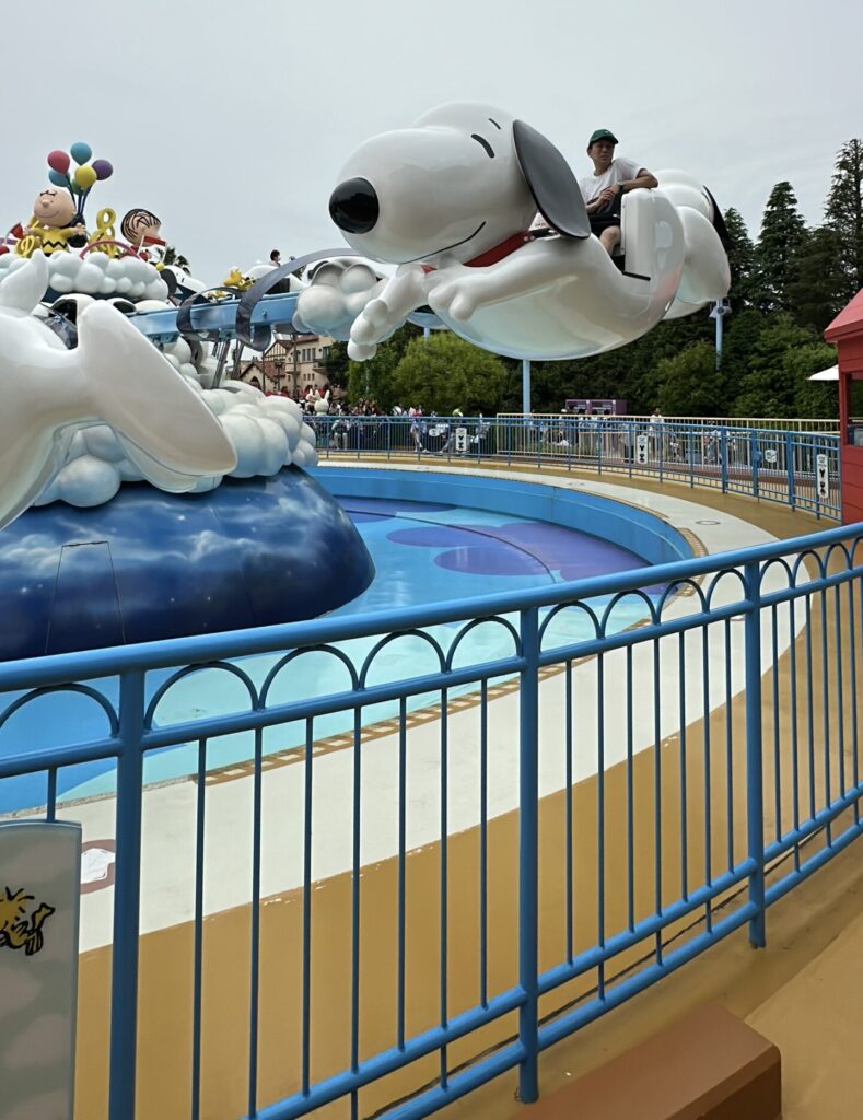 A theme park ride featuring large, cartoonish figures of Snoopy and other Peanuts characters. The ride is designed to look like Snoopy is flying, with people seated in the character's back. The ride is surrounded by a blue fence, and there are trees and buildings in the background.