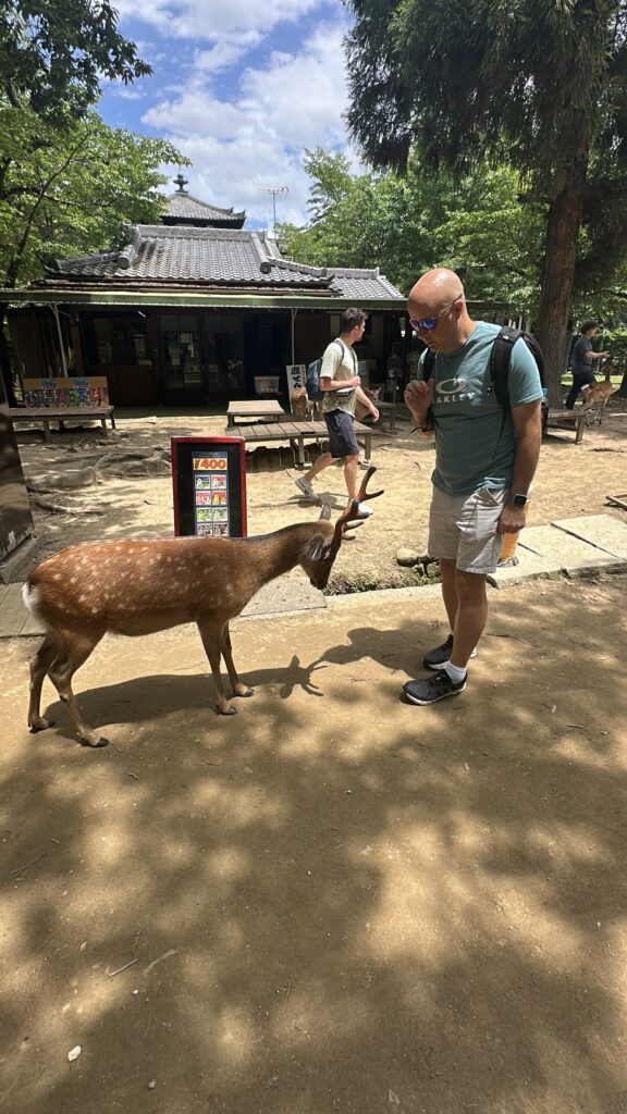 A man wearing sunglasses and a backpack is standing on a dirt path, interacting with a deer. The deer is facing him, and there is a sign with a price in yen nearby. In the background, another person is walking, and there is a traditional building with a tiled roof surrounded by trees. The sky is partly cloudy.