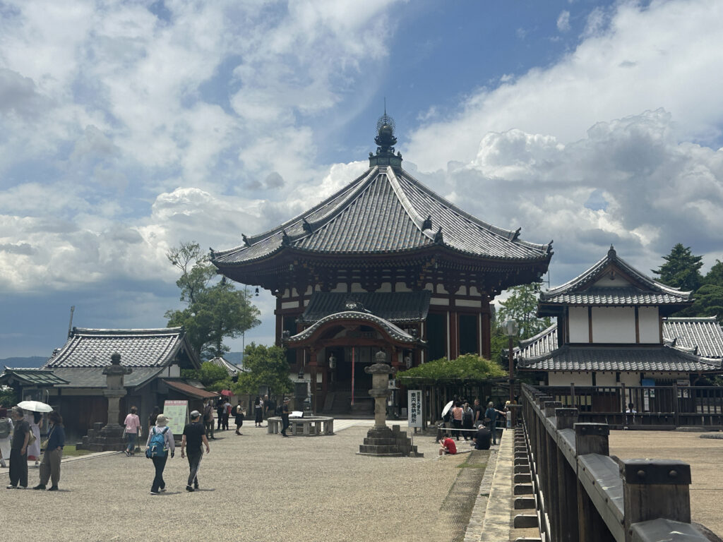 The image shows a traditional Japanese temple complex with several buildings featuring curved, tiled roofs. The main building is prominent in the center, with intricate architectural details. People are walking around the courtyard, and the sky is partly cloudy, creating a bright and serene atmosphere.