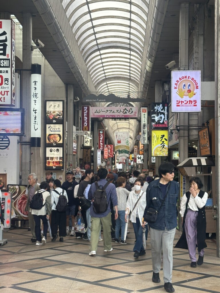 A bustling indoor shopping arcade with a high arched ceiling, featuring numerous signs in Japanese. The area is crowded with people walking in various directions, some carrying umbrellas or wearing masks. The signs advertise different shops and services, including a karaoke room. The atmosphere is lively and busy.