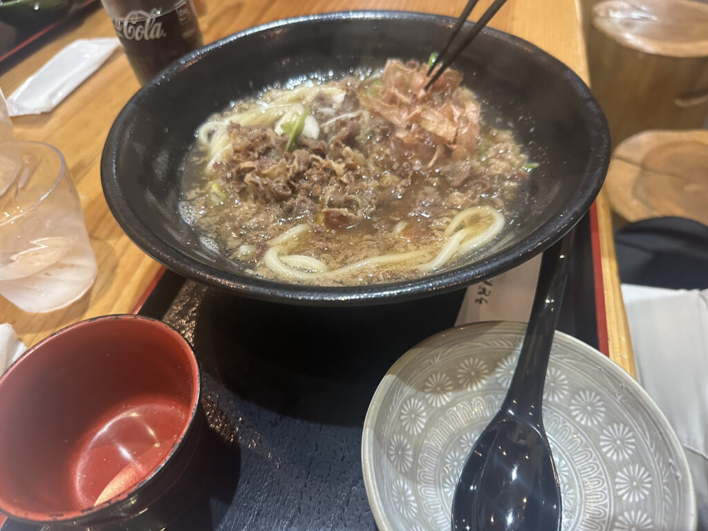 A bowl of noodle soup with slices of meat and garnishes is on a tray. Chopsticks are picking up some of the meat. There is a small empty bowl with a spoon next to it, and a glass of water and a can of Coca-Cola in the background.