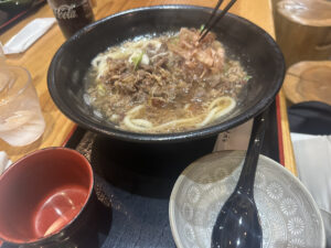 A bowl of noodle soup with slices of meat and garnishes is on a tray. Chopsticks are picking up some of the meat. There is a small empty bowl with a spoon next to it, and a glass of water and a can of Coca-Cola in the background.