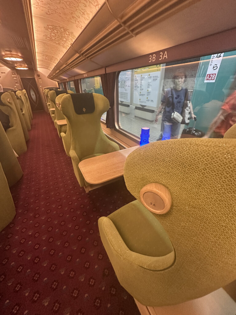 The image shows the interior of a train carriage with plush, green upholstered seats arranged in pairs along a window. Each seat has a small wooden table in front of it. The carpet is red with a patterned design. The ceiling has ornate detailing, and there are overhead lights. Outside the window, a person is visible on the platform, walking past the train. The seat numbers "3A" and "3B" are visible on the headrests.