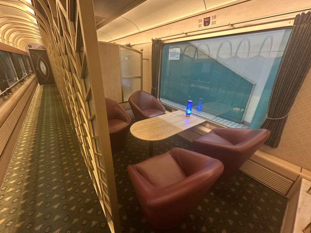 The image shows the interior of a train car with a seating area. There are four red leather chairs arranged around a small wooden table. A blue bottle is placed on the table. The train car has a patterned carpet and a large window with curtains on the right side. The left side features a decorative partition with geometric designs, and the corridor extends towards a door at the end.