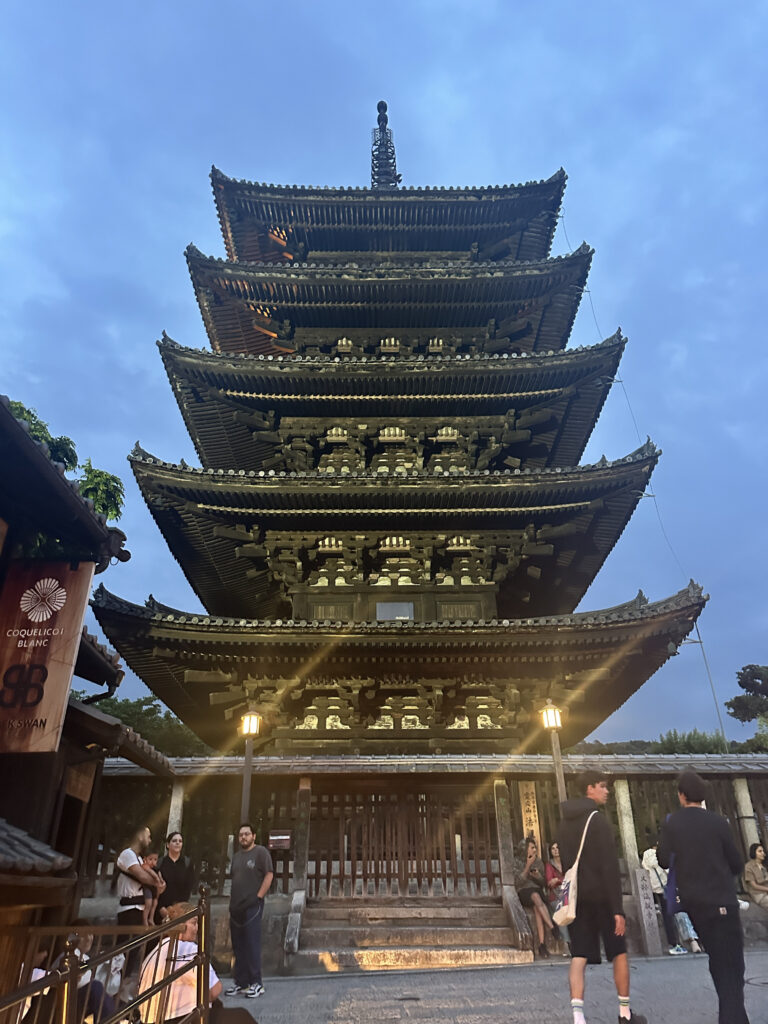 The image shows a traditional multi-tiered pagoda, likely a Japanese temple, with five distinct levels. The structure is ornately designed with intricate wooden details and upturned eaves. It is illuminated by lights, creating a warm glow against the evening sky. Several people are gathered around the base of the pagoda, some standing and others sitting, adding a sense of scale to the impressive structure.