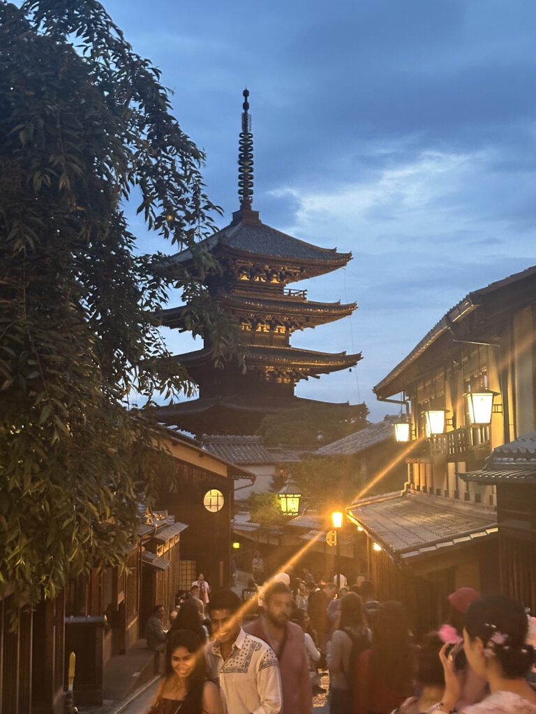 A bustling street scene at dusk in a traditional Japanese area, featuring a prominent pagoda in the background. The street is lined with wooden buildings illuminated by warm lanterns, and many people are walking and enjoying the atmosphere. The sky is a soft blue, adding to the serene ambiance.