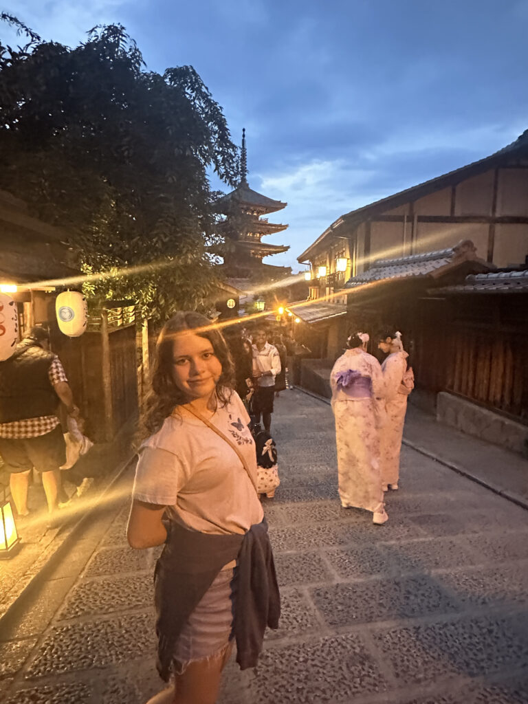 A young woman is standing on a cobblestone street in a historic area during dusk. She is wearing a t-shirt and shorts with a sweater tied around her waist. In the background, there are traditional wooden buildings and a pagoda. Two people wearing kimonos are walking away from the camera. The street is lit with lanterns, creating a warm glow.