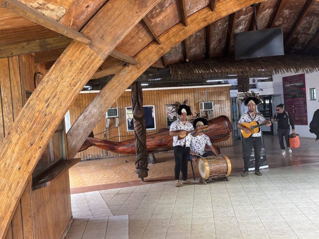 A group of three musicians is performing indoors. They are wearing matching outfits with feathered headdresses. One musician is playing a guitar, another is playing a small stringed instrument, and the third is playing a drum. The setting features wooden architecture with a thatched roof and carved wooden pillars. A person is walking by in the background carrying an orange bag.