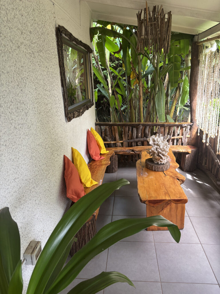 The image shows a cozy outdoor seating area with a rustic wooden bench and table. The bench is adorned with colorful cushions in orange and yellow. A large plant with broad green leaves is in the foreground, and there are more plants in the background, creating a lush, natural atmosphere. A decorative piece made of driftwood hangs from the ceiling, and a mirror with a wooden frame is mounted on the wall. The floor is tiled, and a beaded curtain hangs on the right side.