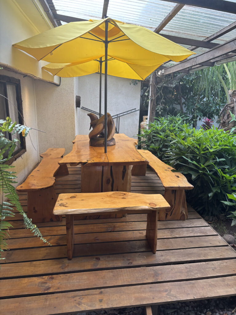 A wooden outdoor seating area with a table and benches is shown. The table has a decorative wooden sculpture in the center and is shaded by a large yellow umbrella. The area is surrounded by greenery, including ferns and other plants, and is situated on a wooden deck next to a building with a window.