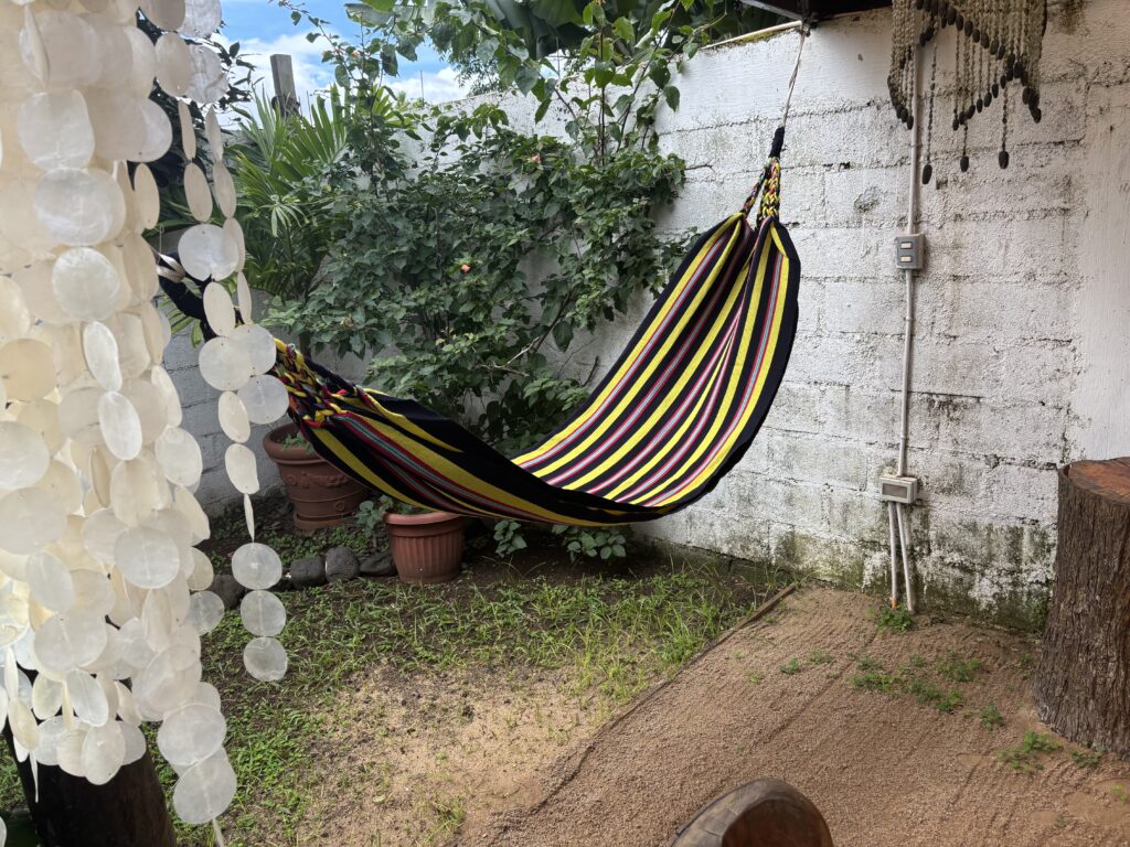 A colorful striped hammock is hanging in a small outdoor area with a white brick wall. There are potted plants and greenery around the hammock. On the left, there are decorative hanging shells, and on the right, a wooden stump is visible. The ground is a mix of grass and sand.