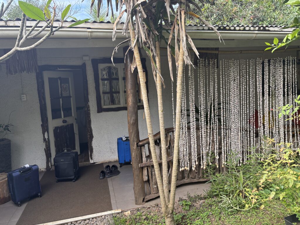 The image shows the entrance to a rustic building with a thatched roof. There are two suitcases and a pair of shoes on a mat in front of the door. The entrance is partially covered by a curtain made of hanging beads. There are plants and trees around the entrance, adding to the natural setting.