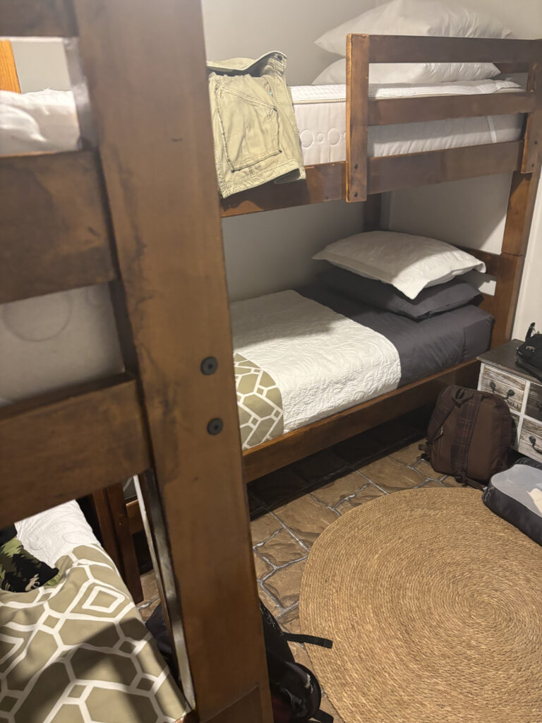The image shows a small room with a wooden bunk bed. The top bunk has a pair of pants draped over the side, and both bunks are made with white and gray bedding. There is a round woven rug on the floor, and a couple of bags are placed nearby. The floor appears to be tiled, and there is a small chest of drawers next to the bed.