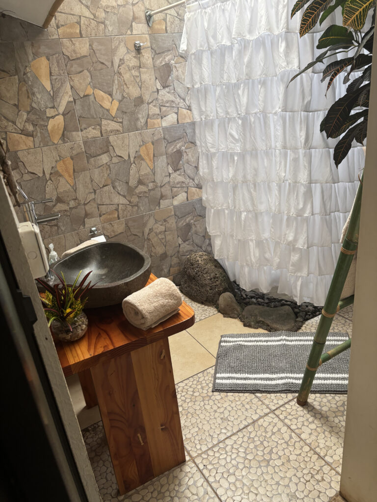 This image shows a bathroom with a rustic and natural design. The walls are covered with stone-patterned tiles. There is a wooden vanity with a stone sink and a rolled-up towel on it. A small plant in a decorative pot is also on the vanity. The shower area is separated by a white, ruffled shower curtain. The floor has pebble-like tiles, and there is a gray and white striped rug. A bamboo ladder and some large stones are visible near the shower area, along with a leafy plant.