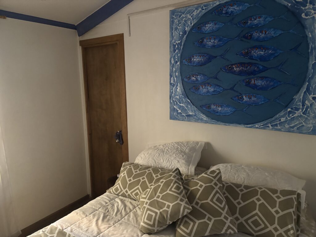 The image shows a bedroom with a bed in the foreground. The bed is covered with a white quilt and has several pillows with geometric patterns in shades of green and white. Above the bed, there is a large painting depicting a circular arrangement of blue fish. The room has a wooden door and white walls with a blue trim near the ceiling.