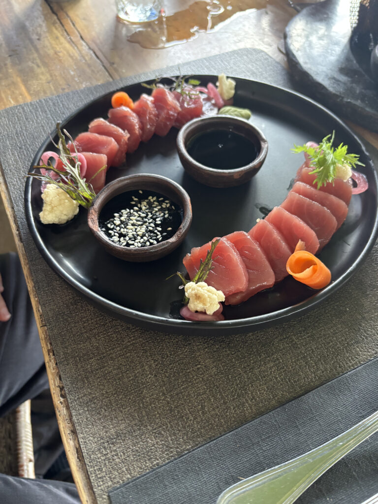A black plate with neatly arranged slices of raw fish, likely tuna, garnished with small vegetable accents like pickled onions, carrot curls, and herbs. There are two small bowls on the plate, one containing soy sauce and the other a sauce topped with sesame seeds. The plate is set on a textured placemat on a wooden table.