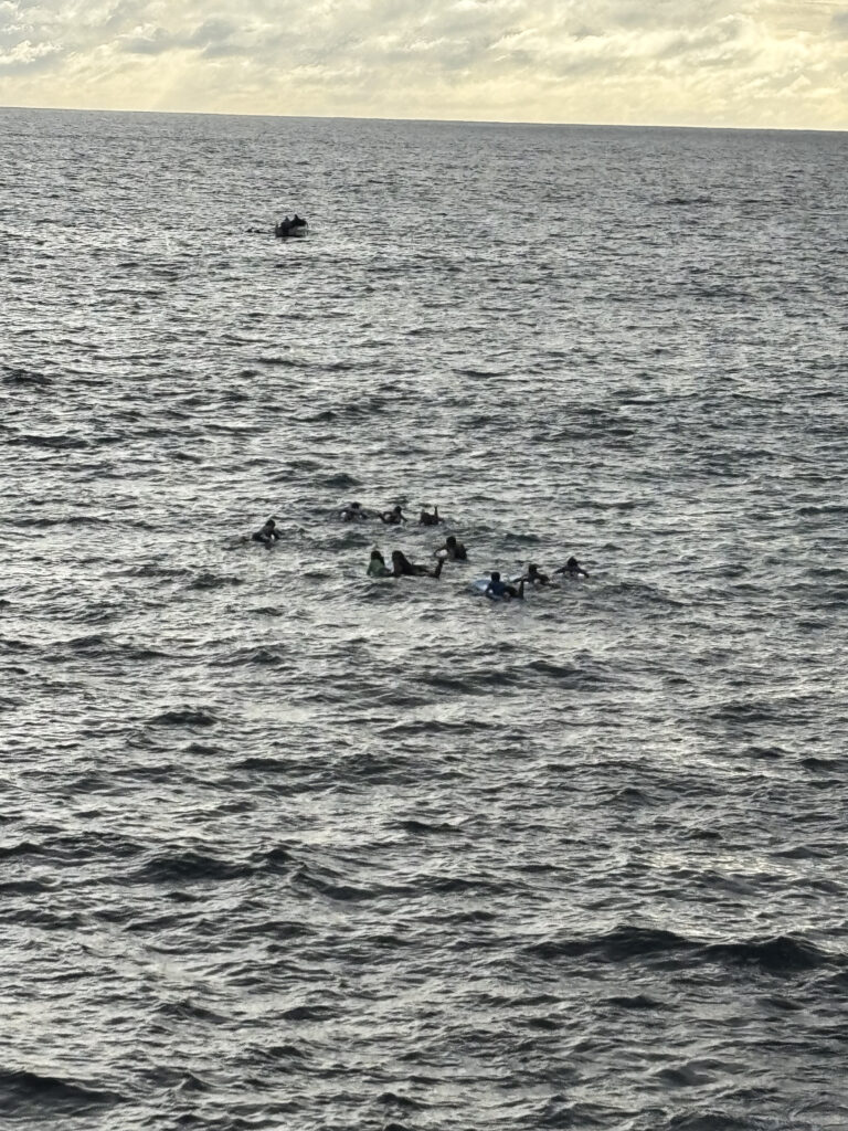 The image shows a group of people in the ocean, possibly swimming or floating together. The water is slightly choppy, and there is a small boat or raft in the distance. The sky is overcast with some light breaking through the clouds.