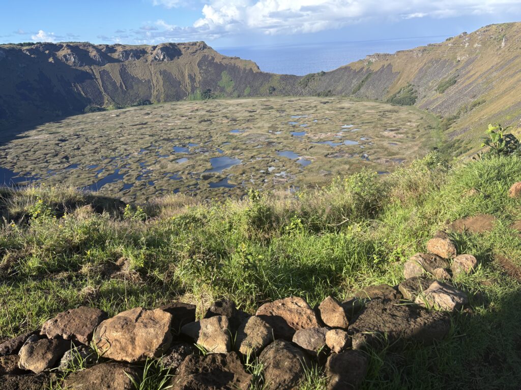 The image shows a large volcanic crater filled with patches of water and vegetation. The crater is surrounded by steep rocky walls, and there is a grassy area with rocks in the foreground. The sky is partly cloudy, and the ocean is visible in the background.