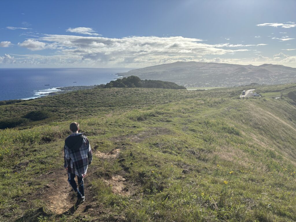 A person is walking along a grassy hilltop path with a scenic view of the ocean and coastline in the background. The sky is partly cloudy, and the sun is shining brightly, casting light over the landscape. In the distance, there are hills and a few scattered buildings.