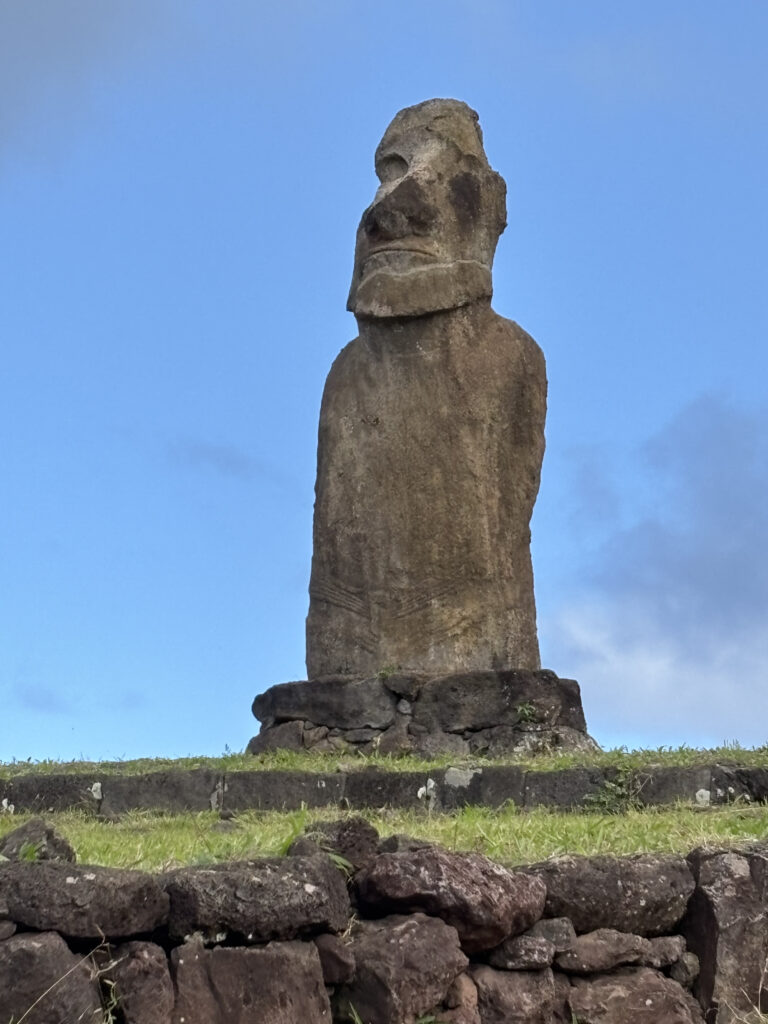 A large stone Moai statue stands on a grassy platform with a stone base. The statue has a prominent head and facial features, typical of the Moai statues found on Easter Island. The sky is clear and blue in the background.