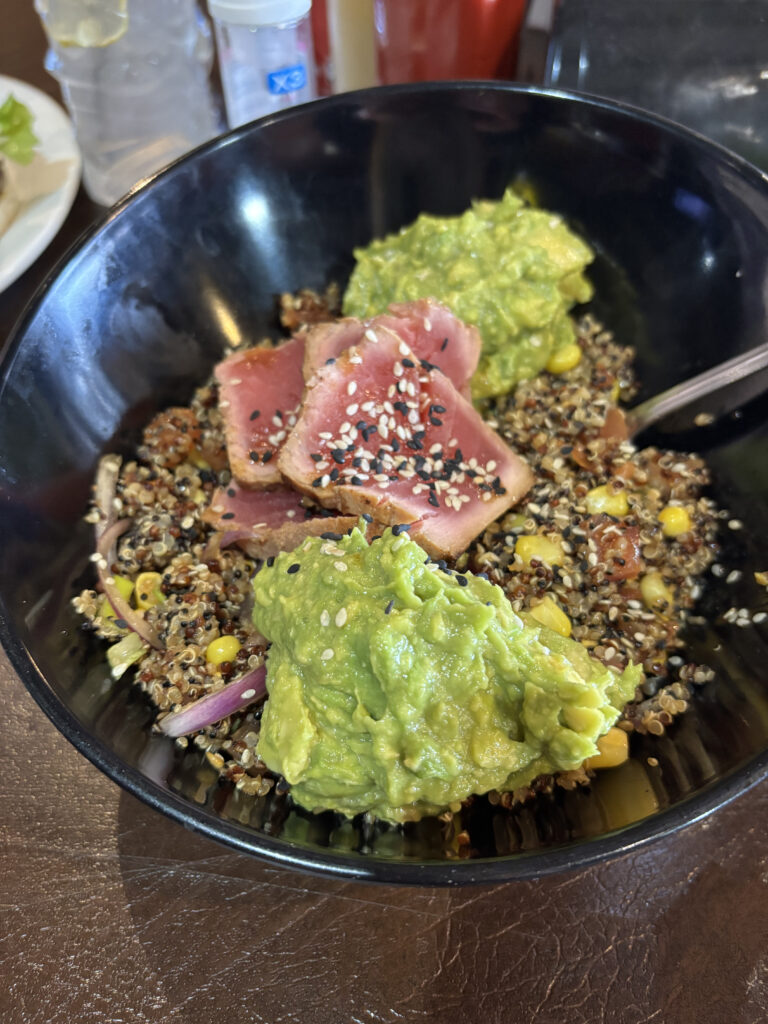 A black bowl containing a dish with sliced seared tuna topped with sesame seeds, a scoop of guacamole, and a quinoa salad with corn and red onions. A fork is placed inside the bowl. In the background, there are condiments and a glass of water with a lemon slice.
