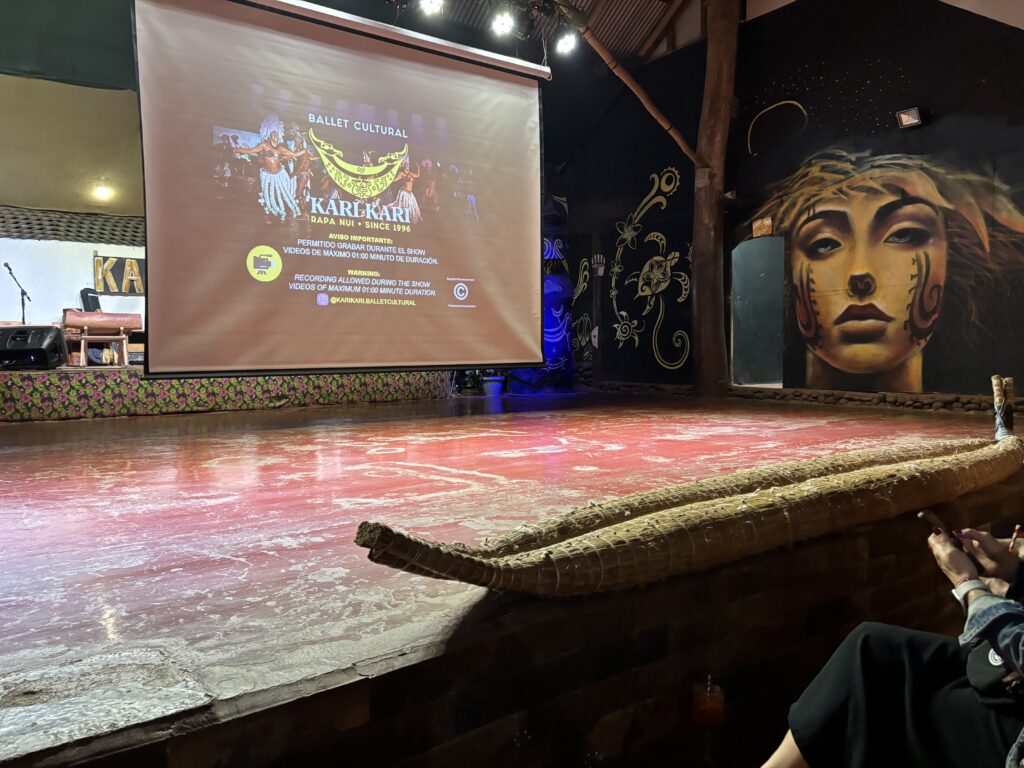 The image shows an indoor stage with a large projection screen displaying information about "Ballet Cultural Kari Kari" from Rapa Nui. The stage has a red floor and is bordered by a woven material. On the right side, there is a mural of a woman's face with traditional face paint. The background includes decorative floral and swirl patterns. In the foreground, a person is seated, partially visible, holding a phone.