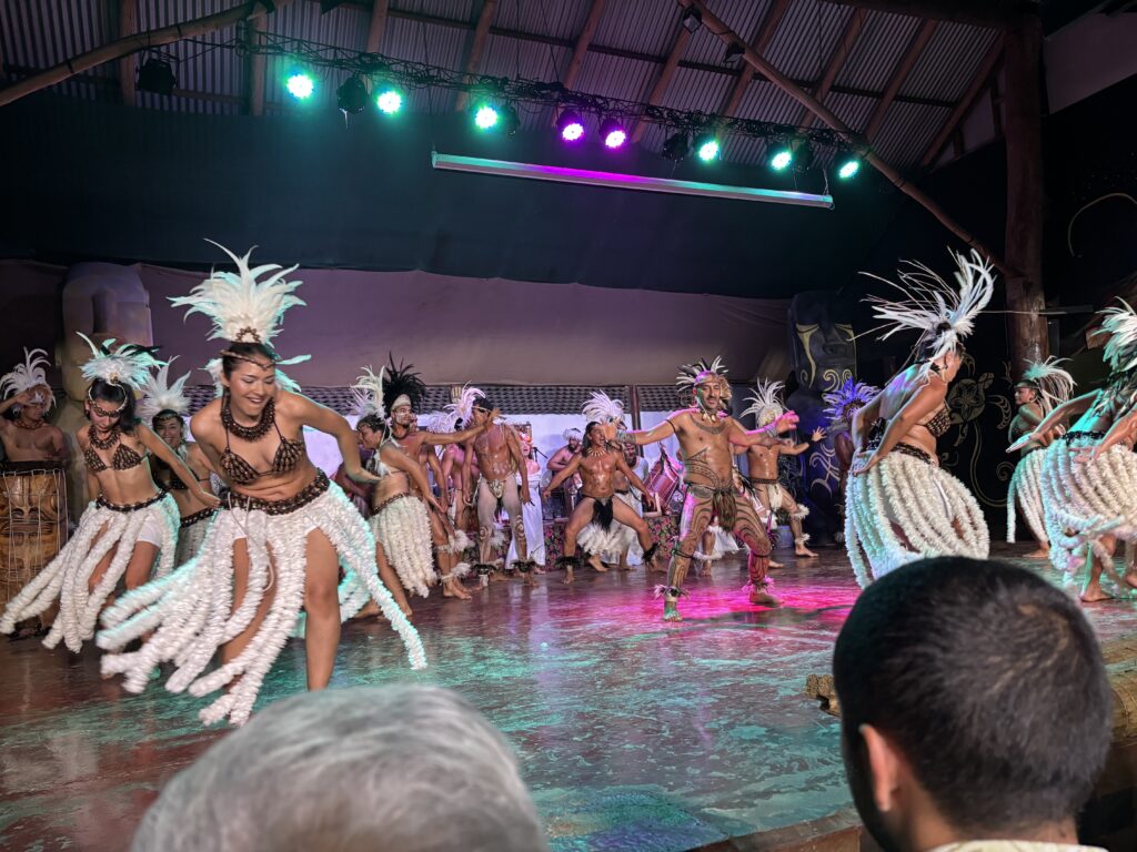 A group of dancers is performing on stage, wearing traditional Polynesian costumes. The women are dressed in feathered skirts and headdresses, while the men have similar attire with decorative body paint. The stage is lit with colorful lights, and the audience is visible in the foreground. The setting appears to be a cultural or traditional dance performance.