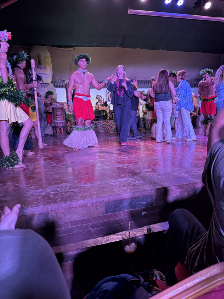 A group of people is on stage participating in a cultural performance. Some are wearing traditional Polynesian attire, including grass skirts and headpieces made of leaves. A woman in the center is holding hands with a performer and smiling. The stage is lit with colorful lights, and there are musical instruments and decorations in the background. Audience members are visible in the foreground, with a drink placed on the edge of the stage.