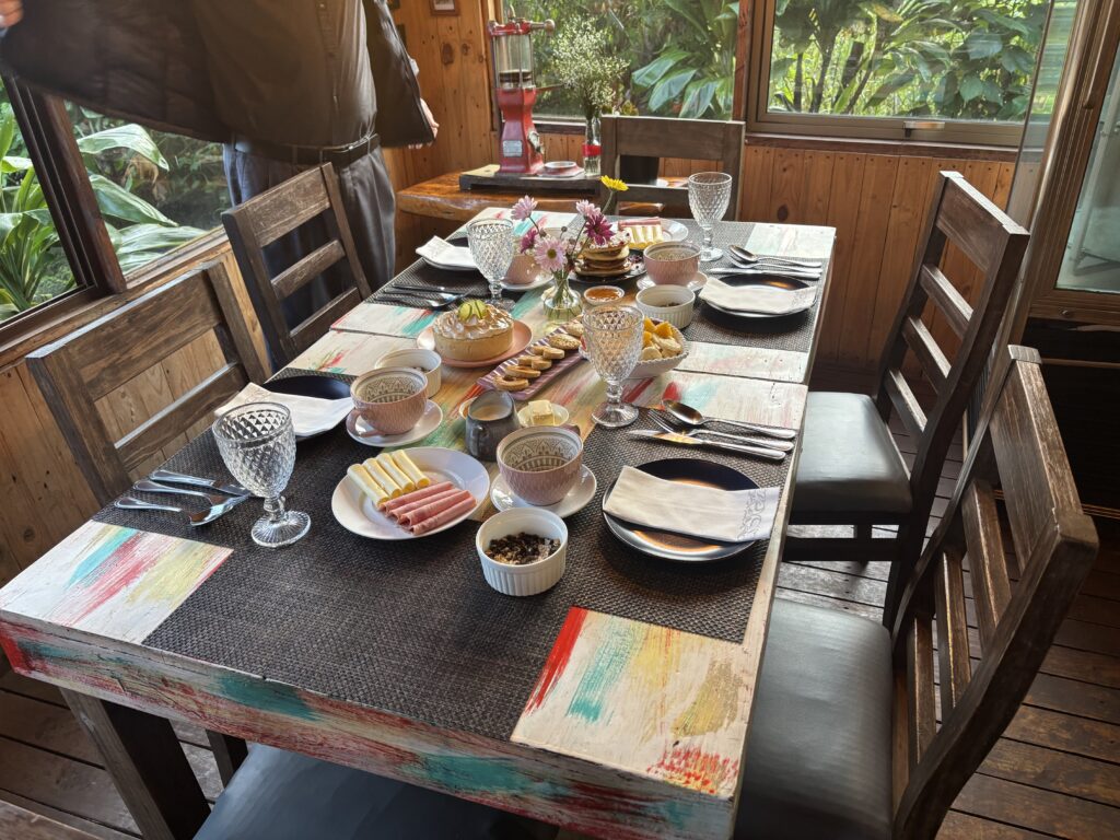 A dining table is set for a meal in a cozy, wooden room with large windows showing greenery outside. The table is decorated with colorful placemats and set with plates, cutlery, and crystal glasses. There are various dishes including rolled cold cuts, cheese, fruit, and small bowls of condiments. A vase with flowers is placed in the center. A person is standing near the window adjusting their jacket.