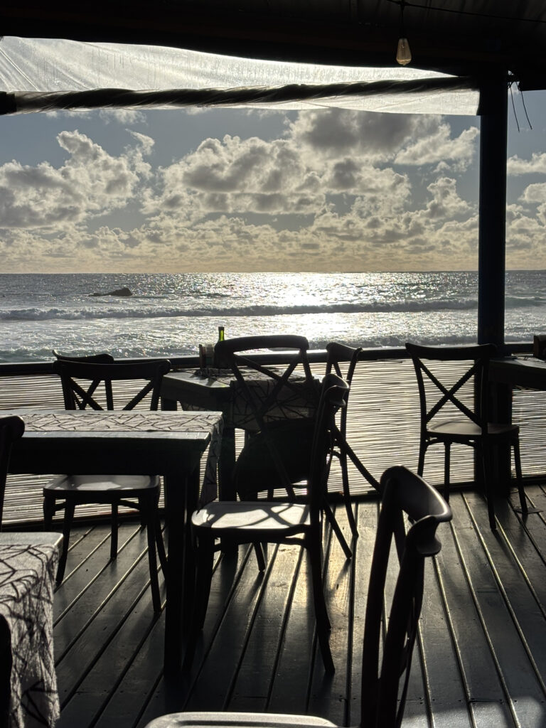 A seaside restaurant with empty tables and chairs on a wooden deck. The sun is setting over the ocean, creating a shimmering reflection on the water. The sky is partly cloudy, and the scene is framed by the restaurant's roof and support beams.