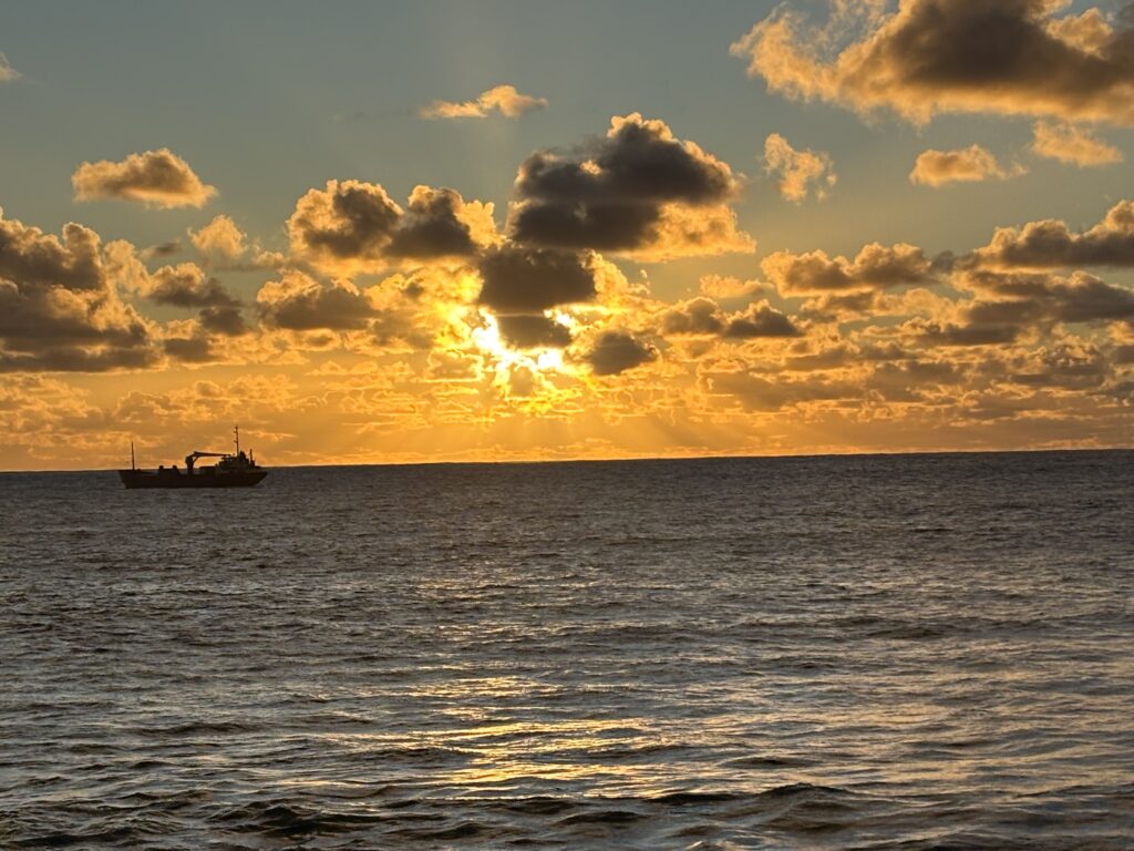 A ship is silhouetted against a vibrant sunset over the ocean. The sky is filled with scattered clouds, illuminated in shades of orange and yellow by the setting sun. The water reflects the warm colors of the sky, creating a serene and picturesque scene.