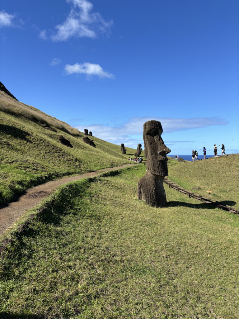 The image shows a landscape on Easter Island with several Moai statues. The statues are large, carved stone figures with distinct facial features, positioned on grassy hills. A dirt path winds through the scene, and a few people are visible in the background, taking photos and walking. The sky is clear and blue with a few clouds.