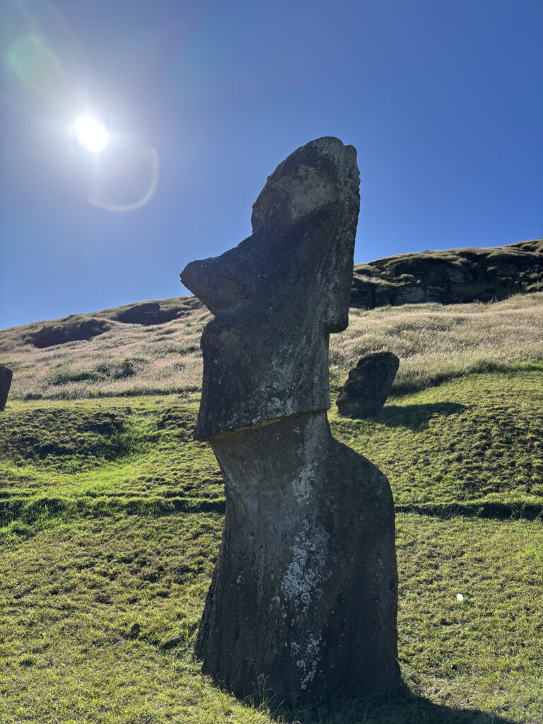 A large stone Moai statue stands on a grassy hillside under a clear blue sky. The sun is shining brightly in the top left corner, casting light and shadows on the landscape.